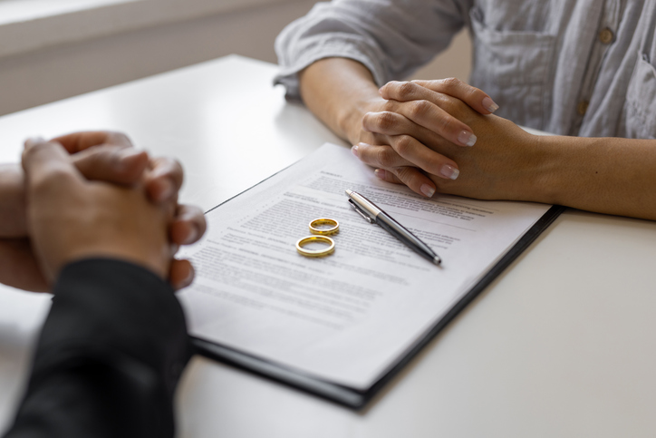 Two people sitting at a table with divorce papers, a pen, and wedding rings placed on top. | Kraft Miles, A Law Corporation